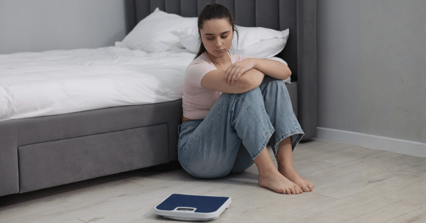 Woman sitting on the floor looking at a scale and feeling frustrated about weight gain and metabolism